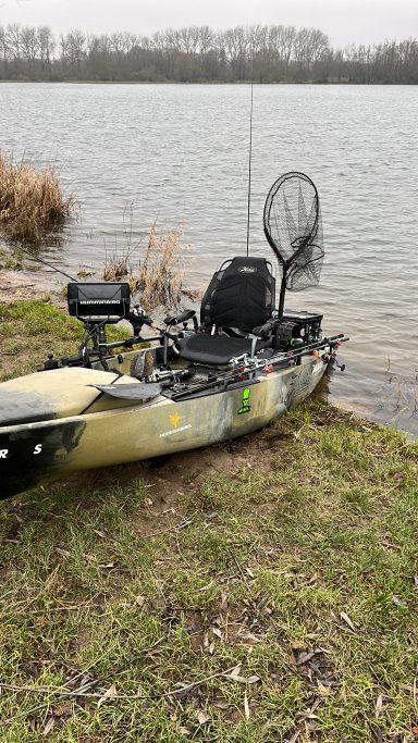 Ein Kayak am Ufer eines Sees, mit einem Netz und Anglerzubehör. Grüne Umgebung.
